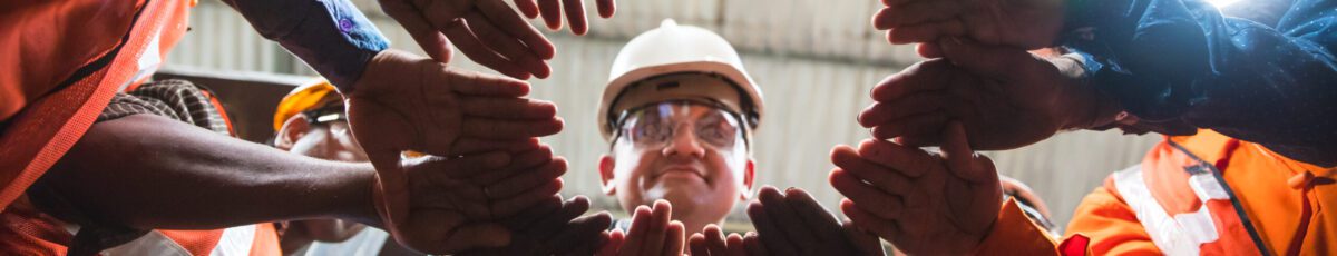 Man looking down towards the camera in the center, with all his factory employees huddled up in a circle with their hands in the center