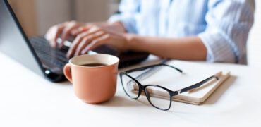 Closeup of a mug of coffee and reading glasses with a person on their laptop in the background.