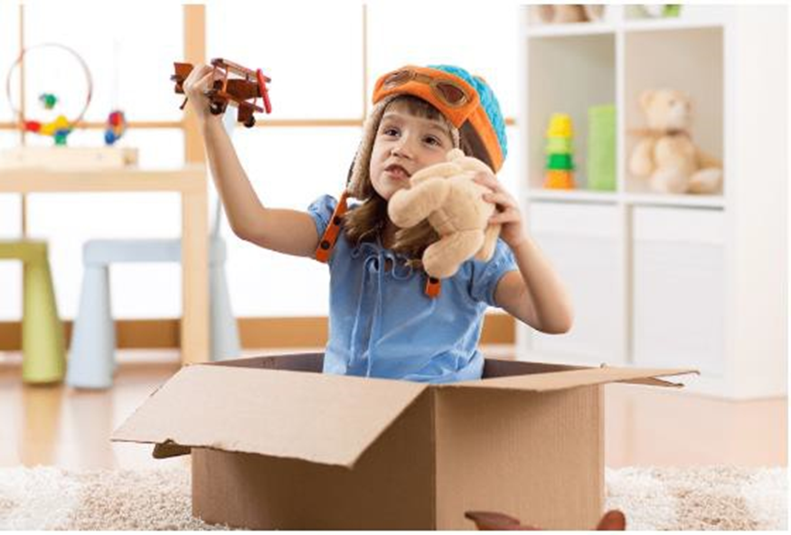 girl sitting inside a carboard box playing with a toy plane and holding a stuffed toy
