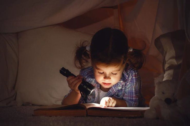 girl with a flashlight reading inside a tent.