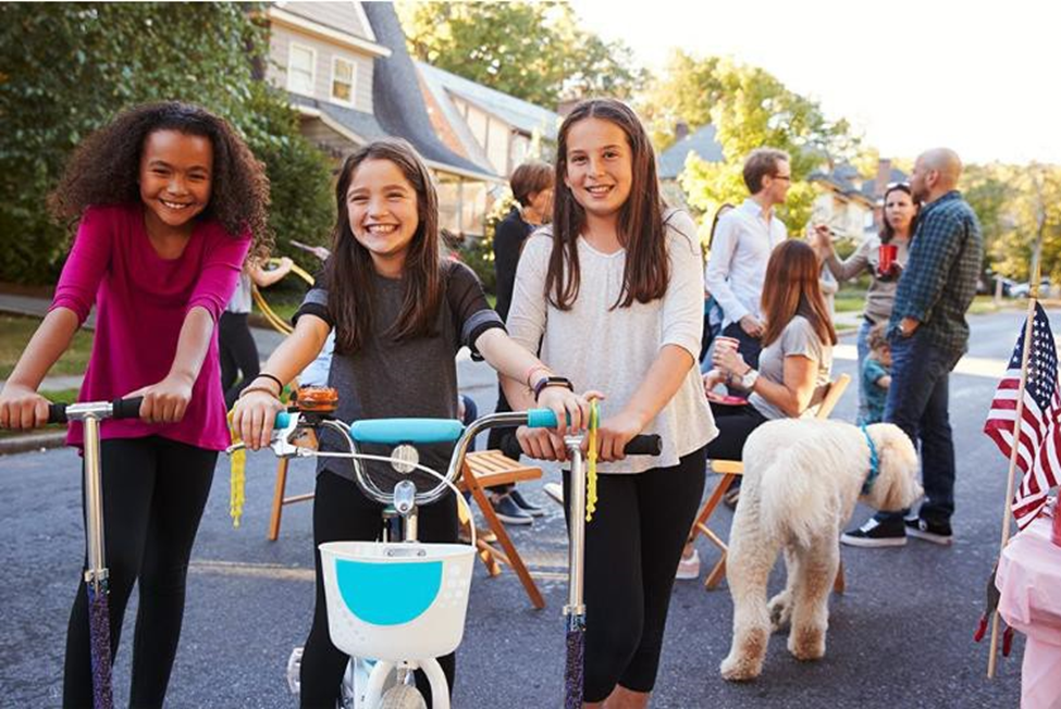 girls riding bike and scooters with their families and neighbors socializing behind them